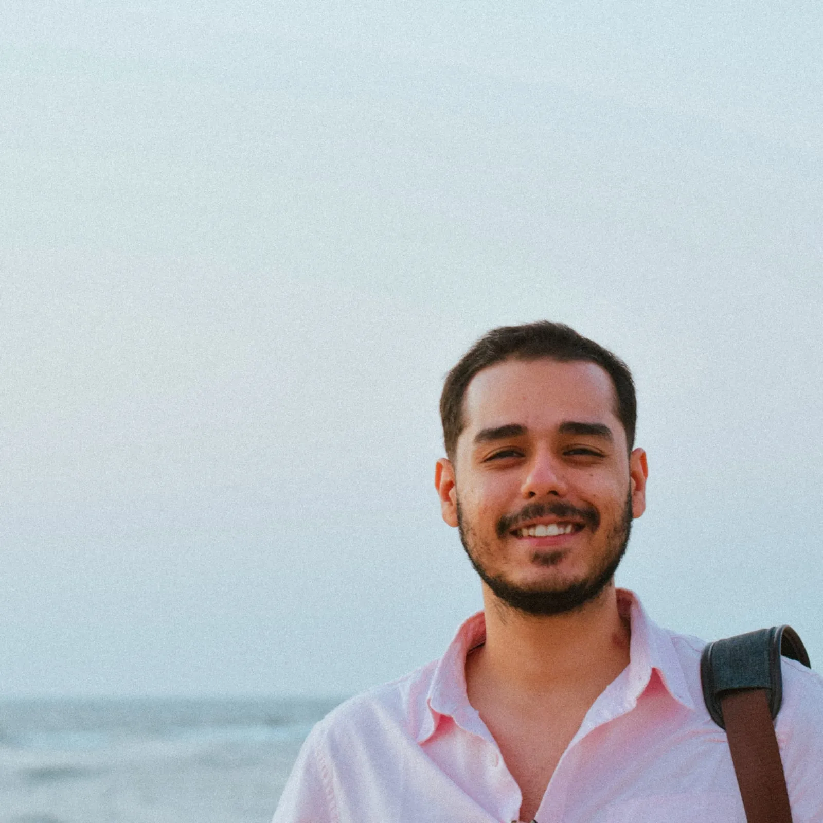 Abhinav smiling on a beach at dusk, wearing a pink shirt with a bag strap over one shoulder
