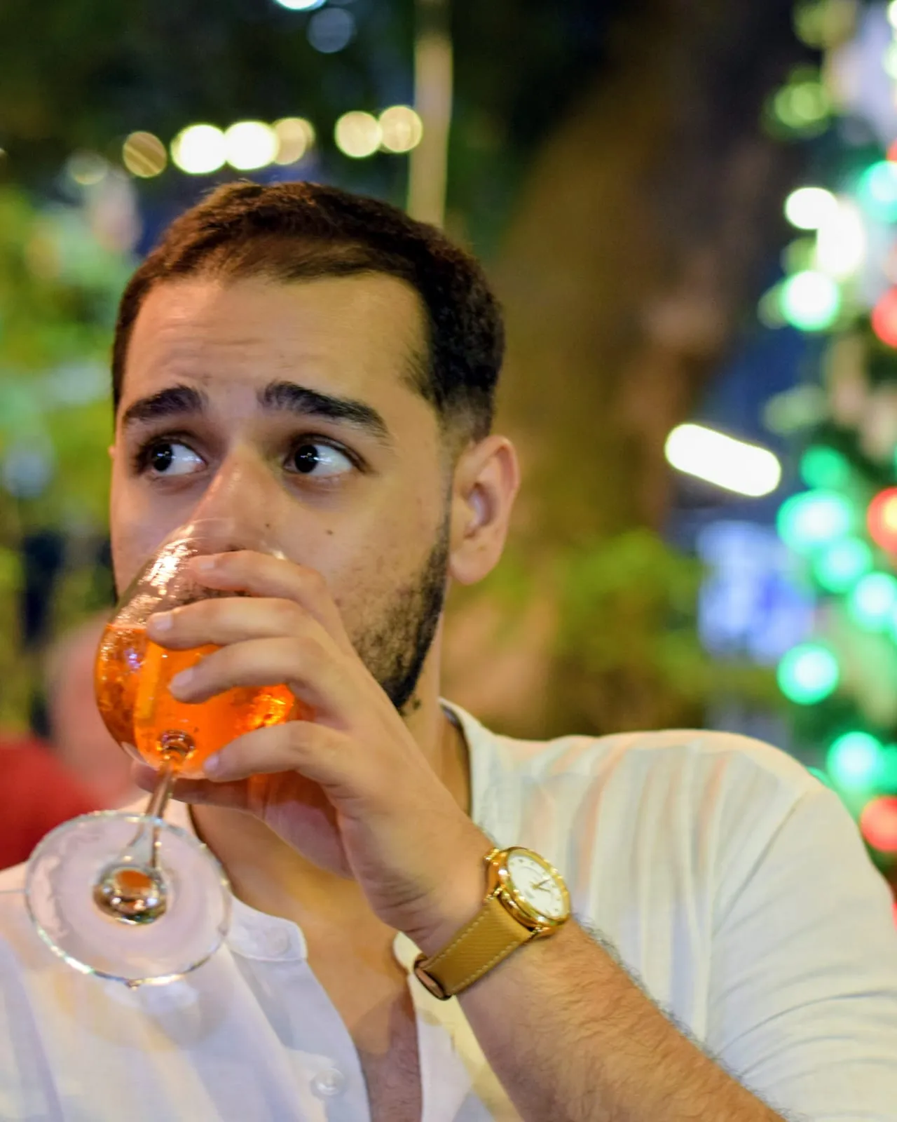 Abhinav sipping an orange drink from a wine glass at an outdoor table lit with string lights