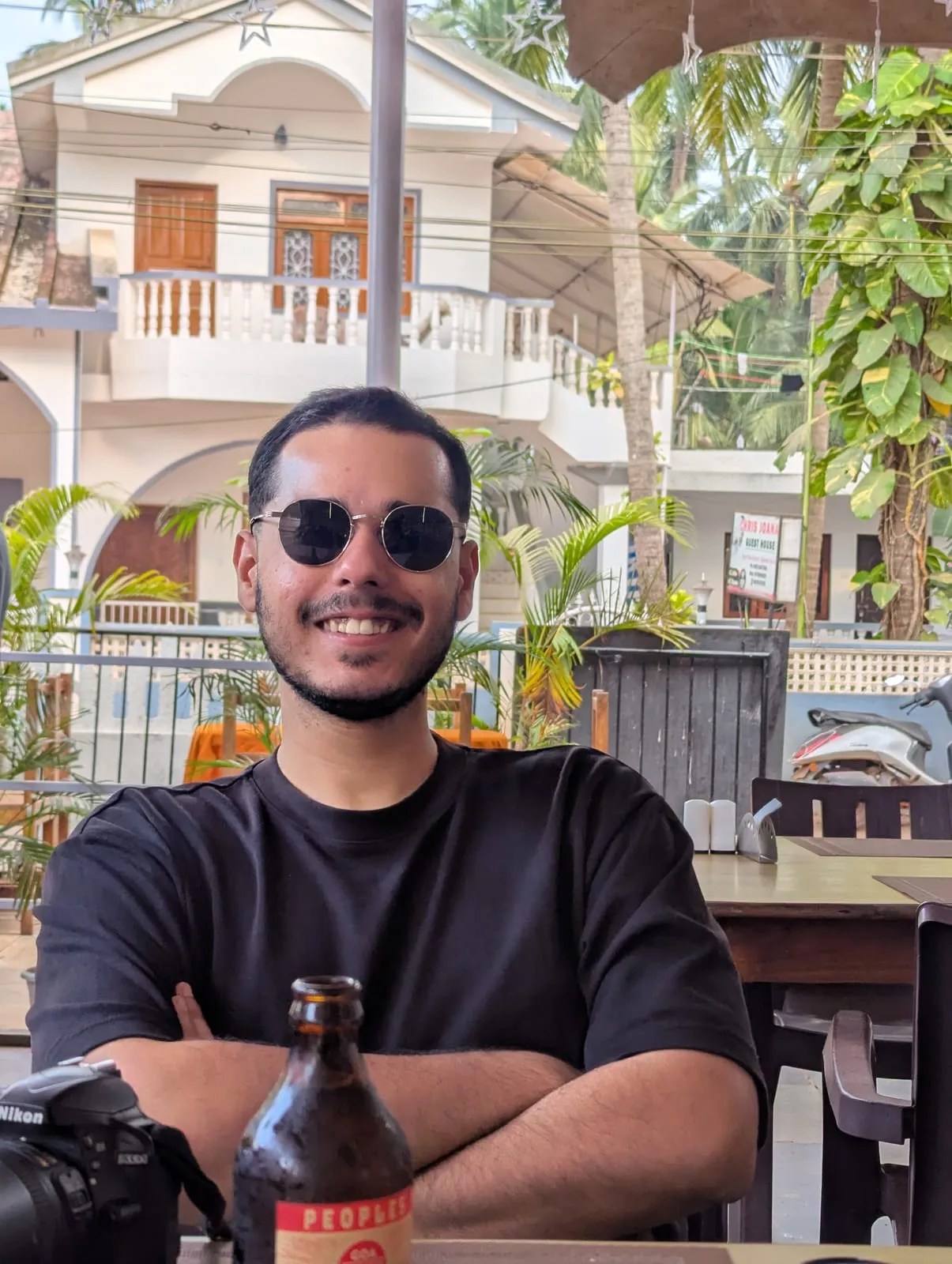 Abhinav at an outdoor café table with a camera and a bottle in front of him, palm trees and a house behind
