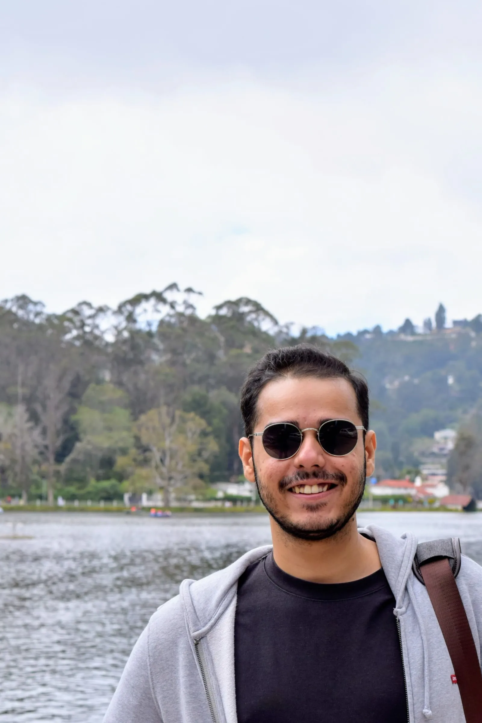 Abhinav grinning in round sunglasses and a grey hoodie, with a lake and a wooded hill behind him