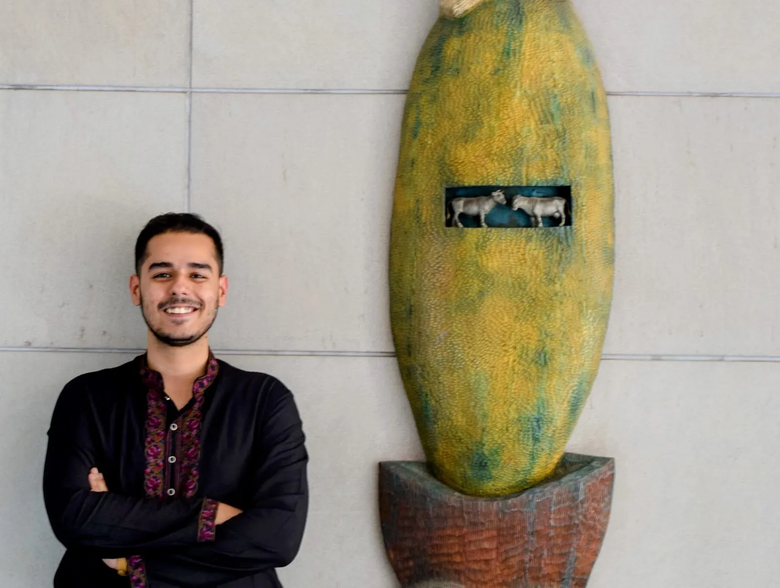 Abhinav smiling beside a tall yellow and green sculpture on a stone wall