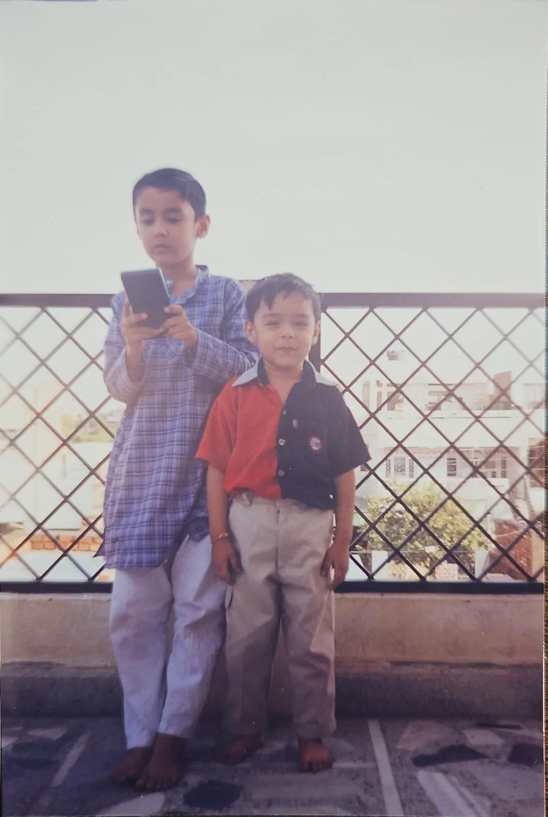 Abhinav as a boy on a terrace, holding up a phone to take a photo, with a younger boy standing beside him